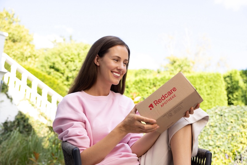 ine lächelnde Frau sitzt im Garten und hält ein Paket von Redcare Apotheke in der Hand.